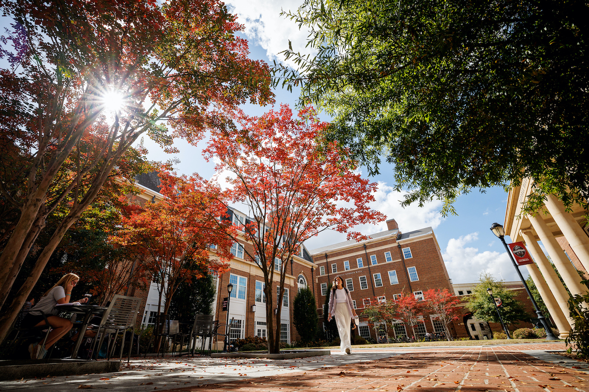 A student walks through a courtyard at the Business Learning Community flanked by fall leaves.