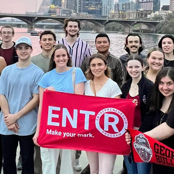 A large group of students stand in front of the Austin Texas skyline holding at UGA Entrepreneurship Program flag