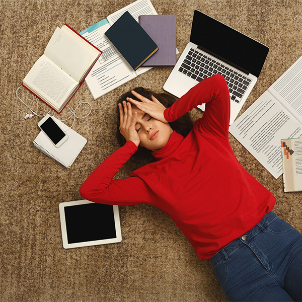 A female high school student lays on the ground and holds her head in distress she is surrounded by a laptop, school books, a phone and papers